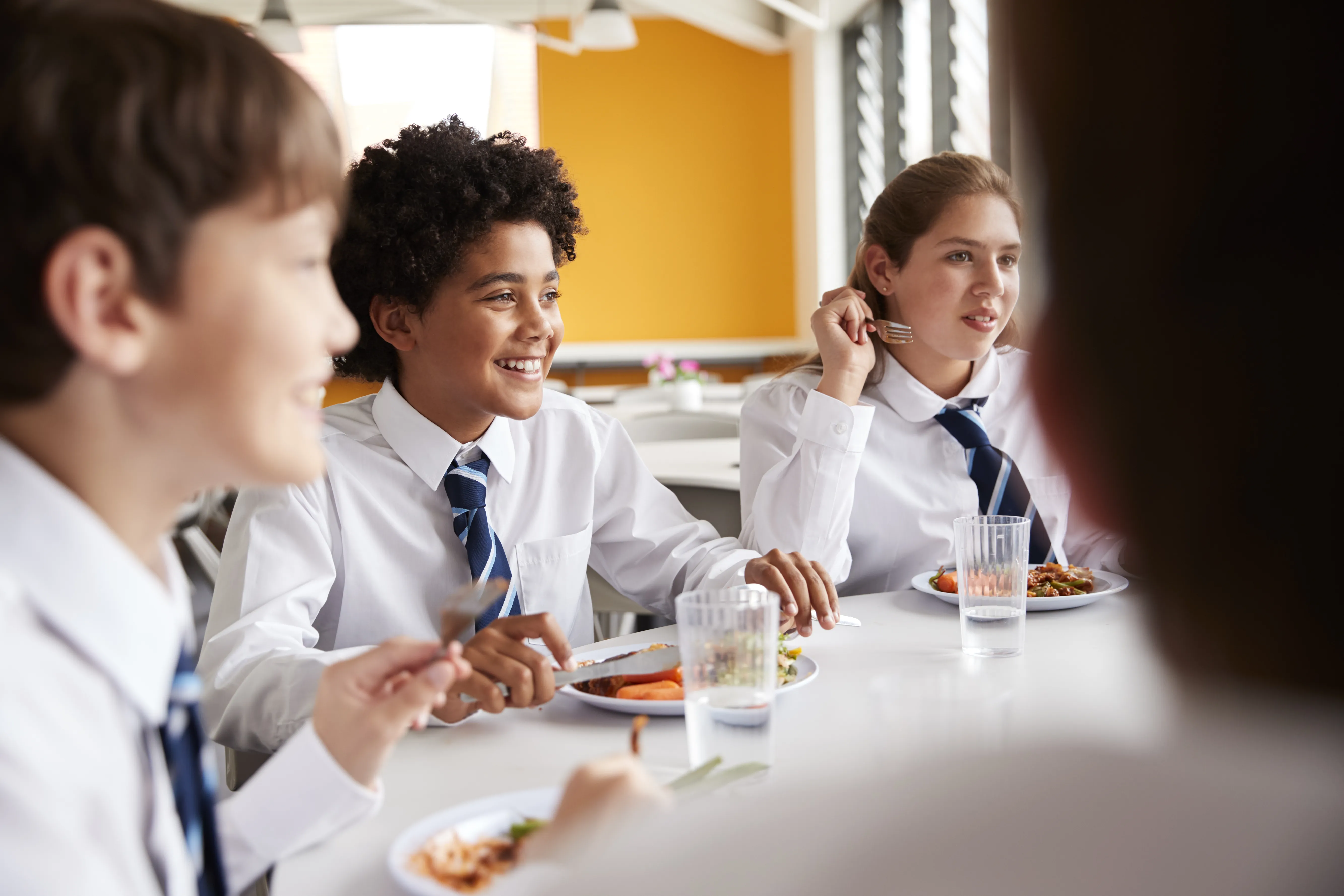 Group Of High School Students Wearing Uniform Sitting Around Table And Eating Lunch In Cafeteria
