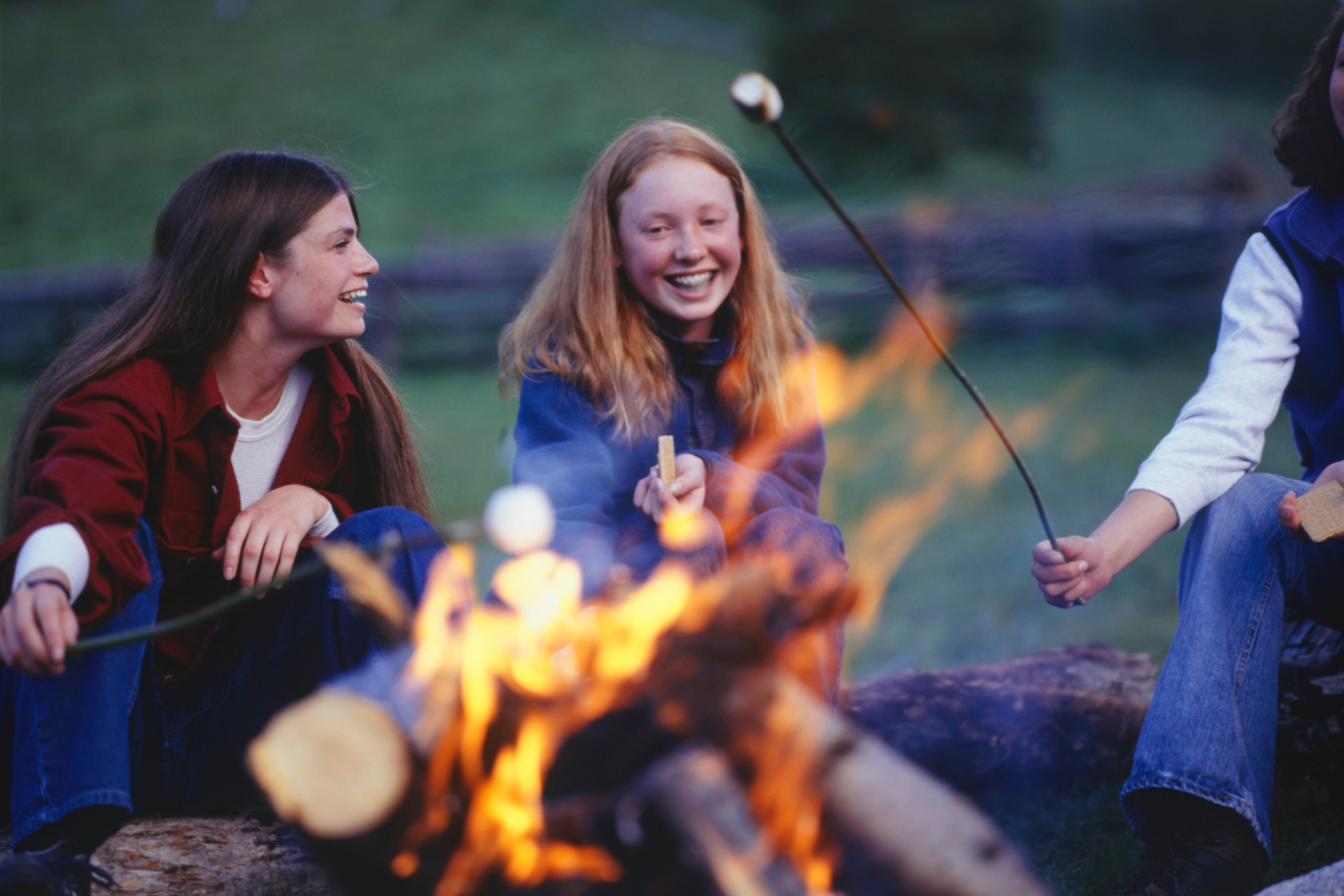 Three teenage girls in country field roasting marshmallows over campfire